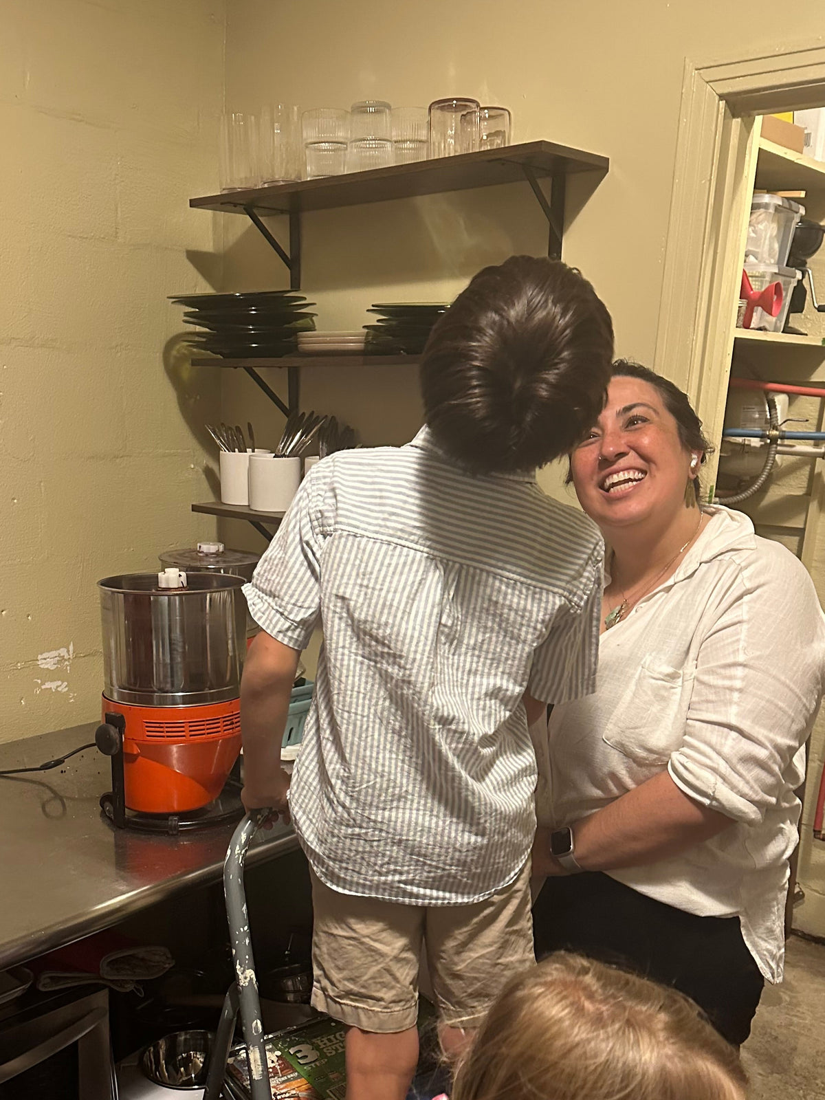A warm, candid kitchen scene featuring founder & chocolate maker Christine and a young boy interacting joyfully while learning about the chocolate making process, and most importantly, tasting the chocolate! Christine is wearing a white long-sleeved top with rolled-up sleeves and black pants, and the child is wearing a white and gray striped short-sleeve shirt and tan shorts. The child is standing on a stepladder to be able to see over the counter.
