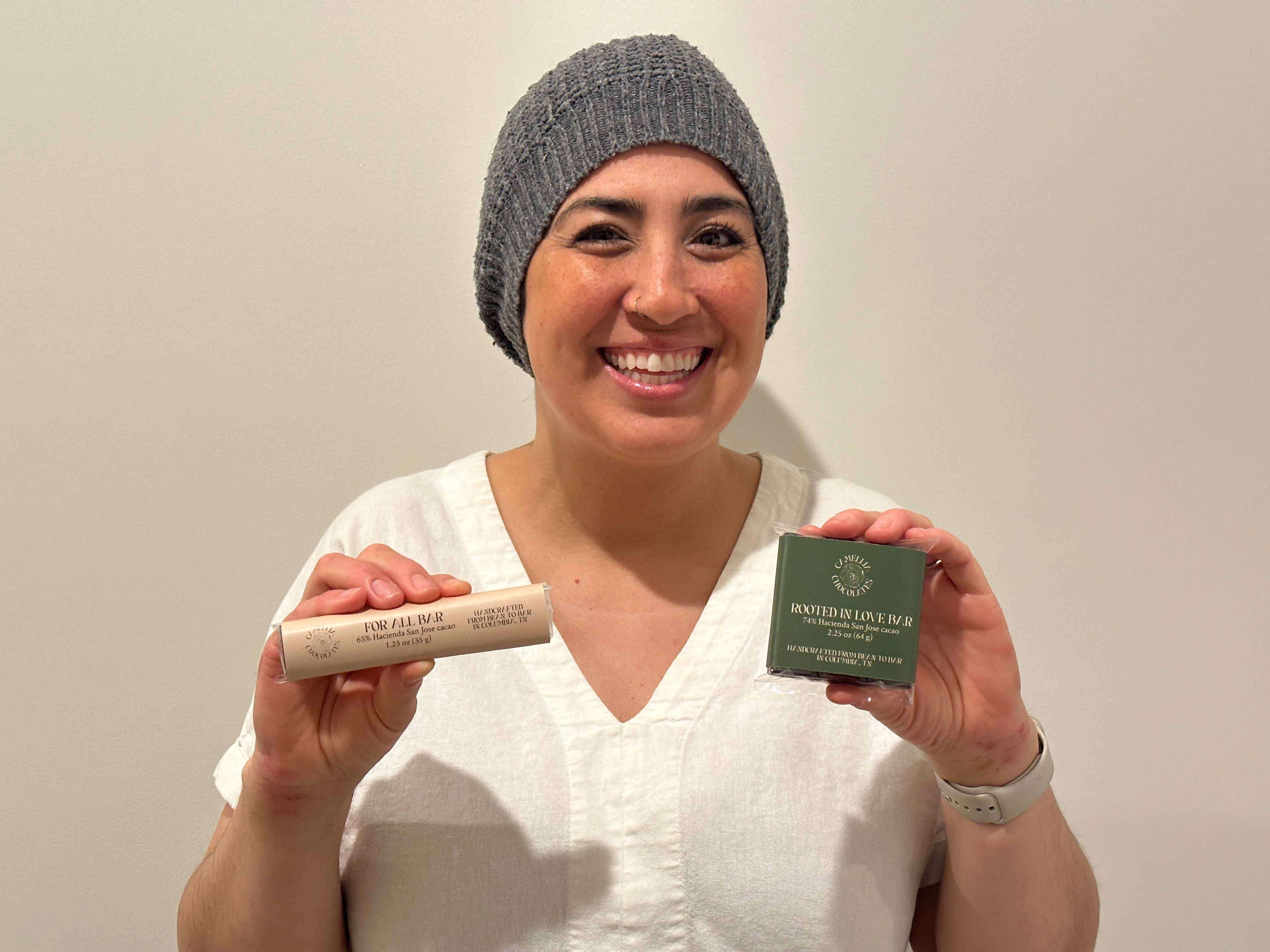 Picture of Camellia Chocolates founder Christine Cole in a white shirt and grey beanie, holding a mini For All Bar in her right hand and a square Rooted in Love Bar in her left hand.
