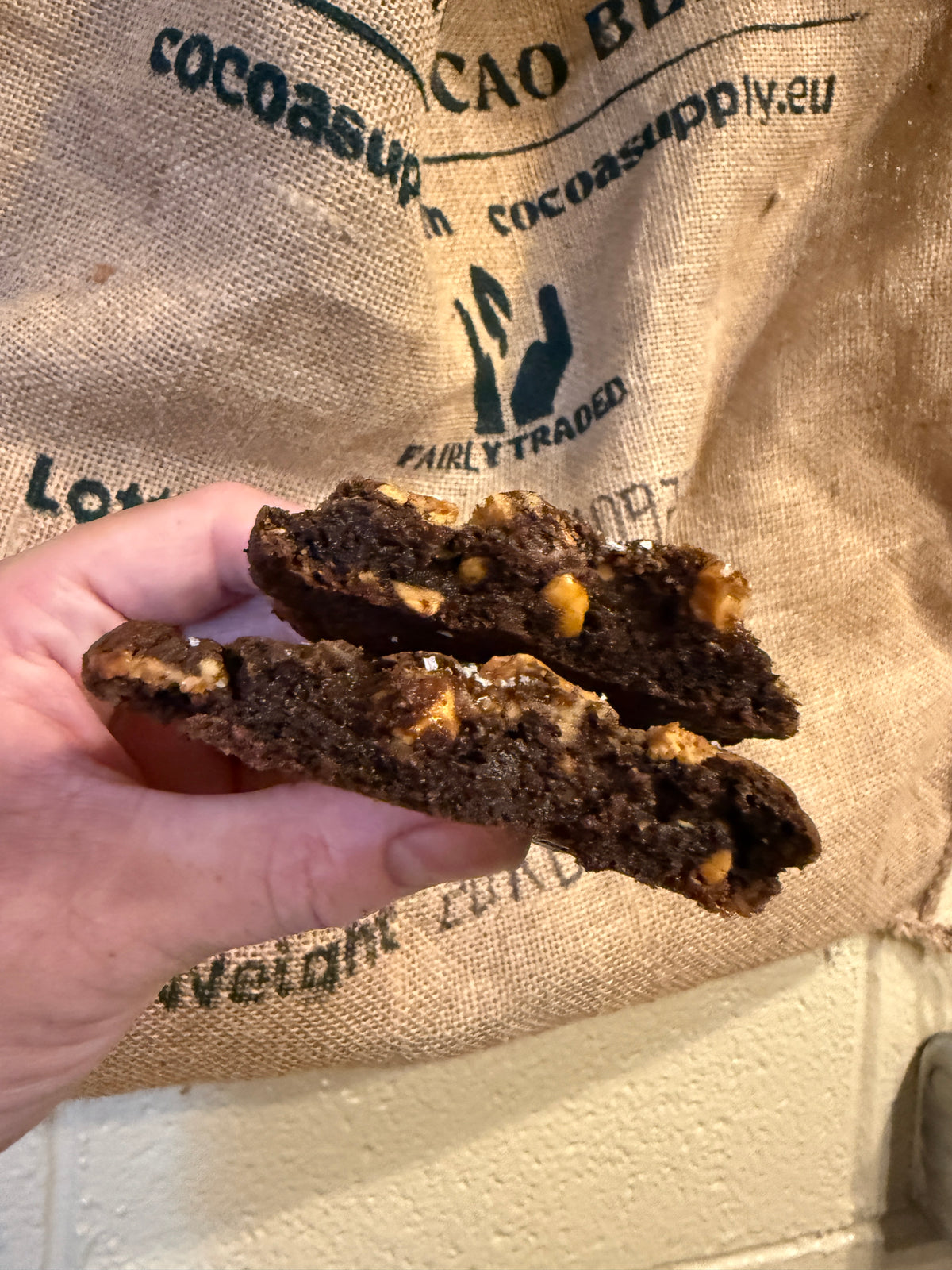 A hand holding two halves of a chocolate cookie with peanut brittle and peanut butter swirl, set against a burlap sack printed with cacao company text.